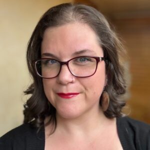 mid-30s woman with dark hair, black-rimmed glasses, and red lipstick